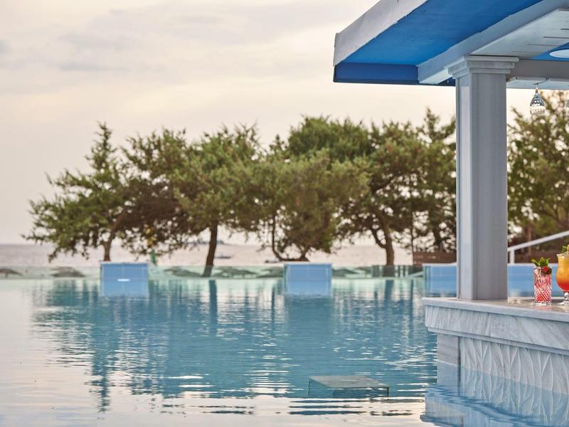 Piscina a sfioro con bar in piscina, vista su alberi e mare al tramonto.