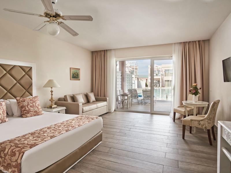 Camera d'hotel luminosa con letto matrimoniale, zona soggiorno e balcone con vista sulla piscina.