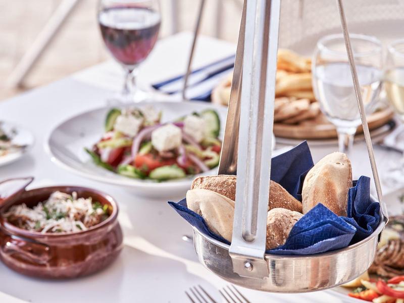 Tavolo apparecchiato con piatti mediterranei, cesta di pane e bicchieri di vino su tovaglia bianca.