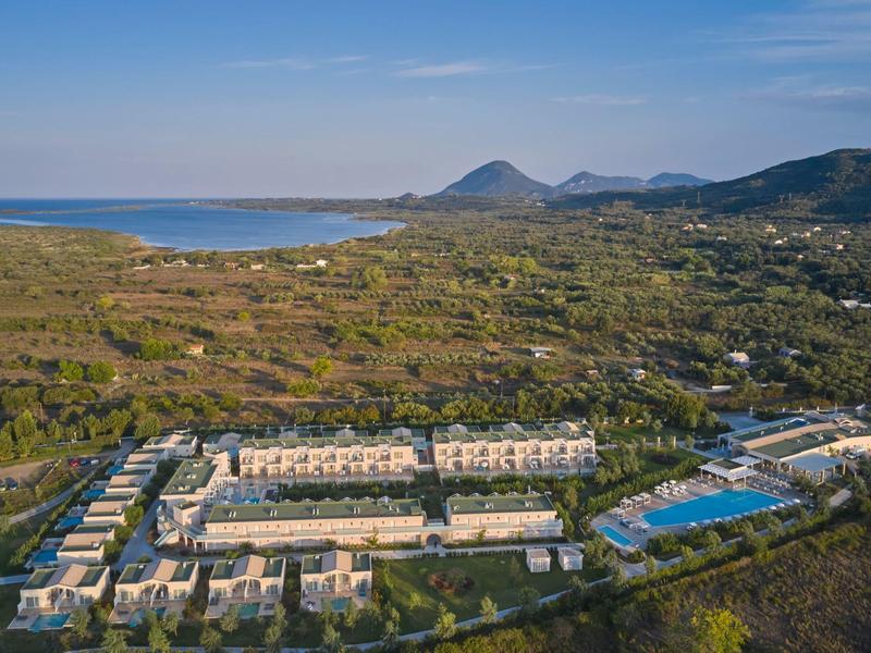 Veduta aerea di un resort con piscine, circondato da montagne e mare sullo sfondo.