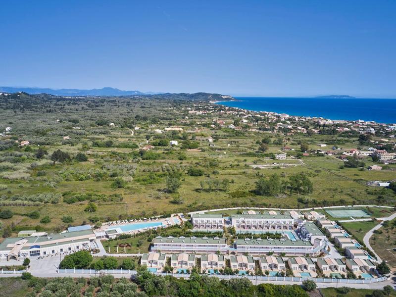 Veduta aerea di un paesaggio costiero con case residenziali e vegetazione sotto un cielo azzurro.