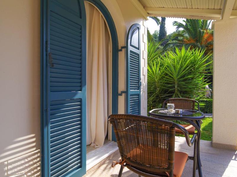 Veranda mit blauem Fensterladen, Holztisch, Korbstuhl und grüner Gartenpflanze im Hintergrund.