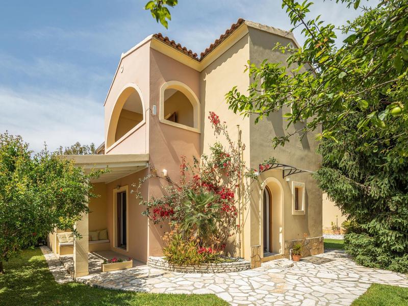 Haus mit hellbeiger Fassade, roten Blumen, Bäumen und weißem Steinweg im Garten bei blauem Himmel.