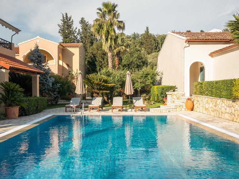 Ein klarer Pool umgeben von mediterranen Häusern, Palmen und Sonnenliegen unter blauem Himmel.