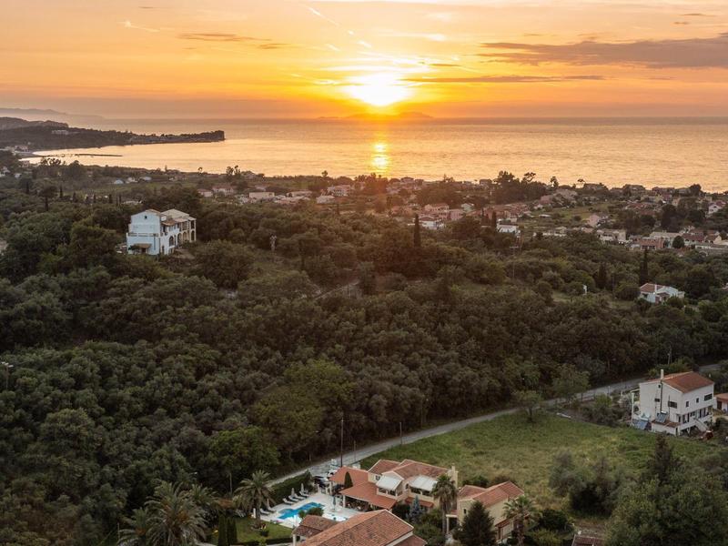 Sonnenuntergang über grünem Wald und mehreren Häusern nahe der Küste mit ruhigem Meer.