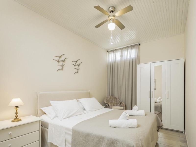 Helles Schlafzimmer mit Doppelbett, Nachttisch, grauen Vorhängen, Kleiderschrank und Deckenventilator.