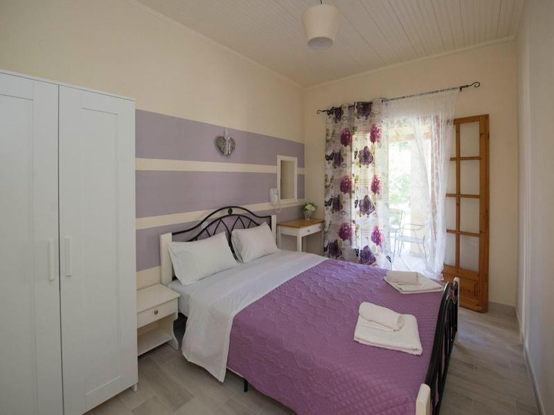 Schlafzimmer mit lila Bettdecke, weißen Möbeln und Blumen- Gardinen am Fenster.