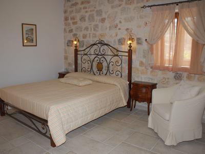 Schlafzimmer mit Steinmauer, Metallbett, cremefarbener Bettdecke, Nachttischen und weißem Sessel.