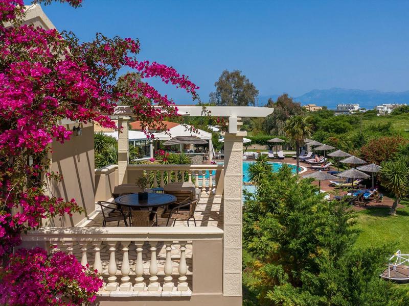 Balkon met tafels en stoelen met uitzicht op een tuin met zwembad en ligstoelen onder een heldere hemel.