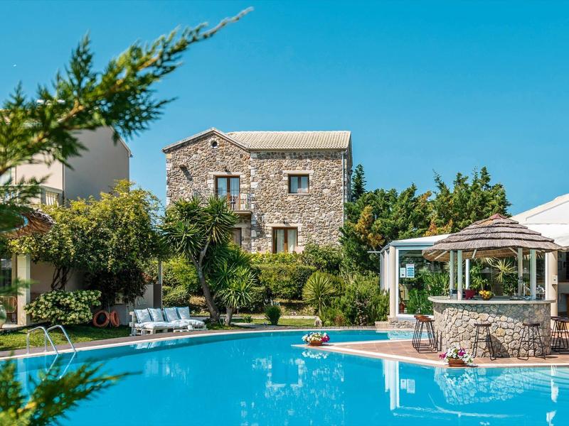 Hotel met stenen muren en groot zwembad omgeven door groene bomen en parasols.