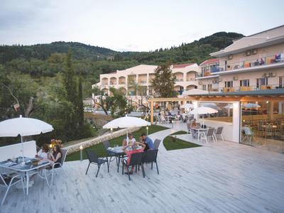 Hotel mit Terrasse, Tischen, Stühlen, Sonnenschirm und grünen Hügeln im Hintergrund bei bewölktem Himmel.