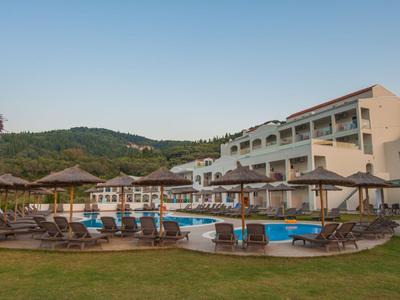 Hôtel avec piscine, chaises longues et parasols devant des collines boisées sous un ciel dégagé.