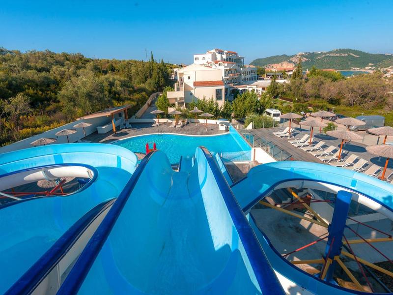 Widok z góry na niebieskie zjeżdżalnie wodne na hotelowym basenie z leżakami i wzgórzami w tle.