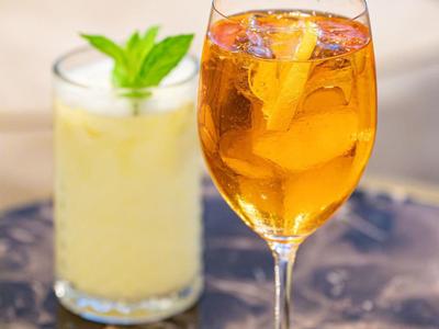 Een gouden cocktail met ijs en sinaasappelschil naast een romige drank met munt op marmeren tafel.