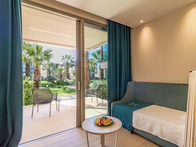 Hotelkamer met slaapbank, kleine tafel en uitzicht op tuin terras met stoelen buiten.