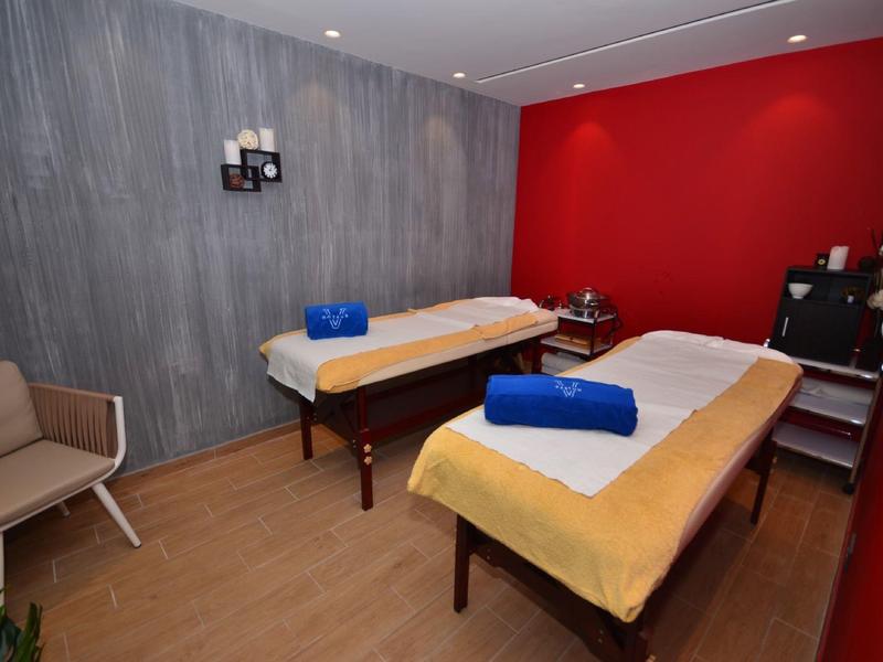 Gezellige massageruimte met twee behandelbedden, handdoeken, een stoel en rode en grijze muren.