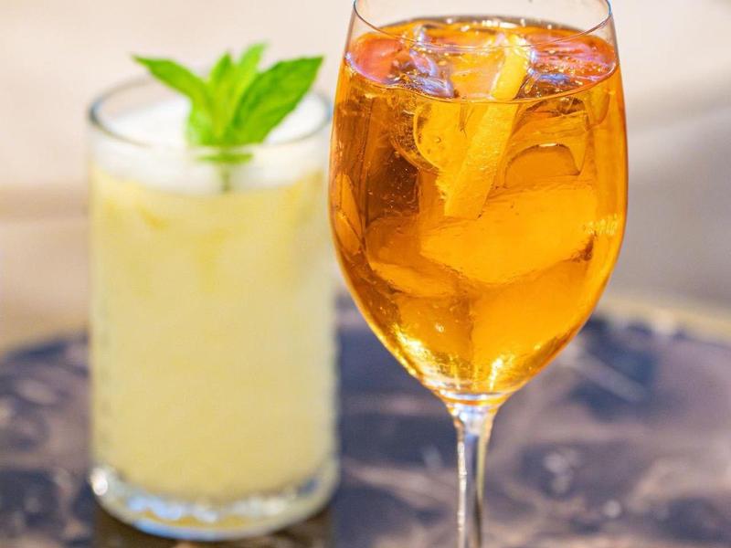 Een gouden cocktail met ijs en sinaasappelschil naast een romige drank met munt op marmeren tafel.