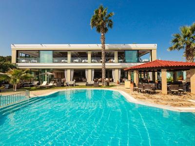 Moderner zweistöckiger Gebäudekomplex mit großem Pool, Palmen und Sonnenterrasse unter blauem Himmel.