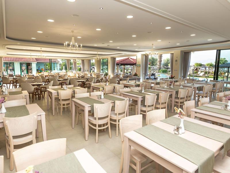 Großer heller Speisesaal mit vielen weißen Holztischen und Stühlen, großen Fenstern und grünen Tischläufern.