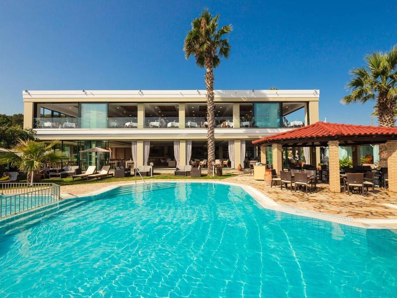 Moderner zweistöckiger Gebäudekomplex mit großem Pool, Palmen und Sonnenterrasse unter blauem Himmel.