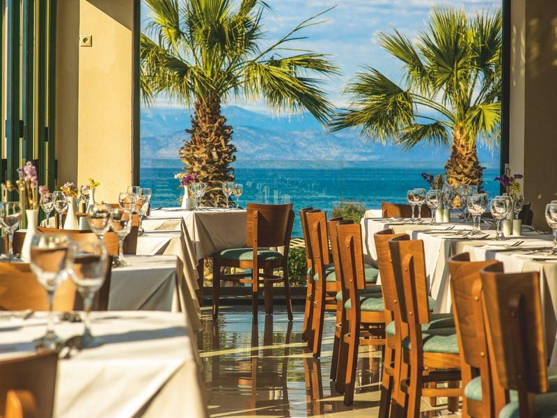 Offenes Restaurant mit Holztischen, weißen Tischdecken und Meerblick unter Palmen.