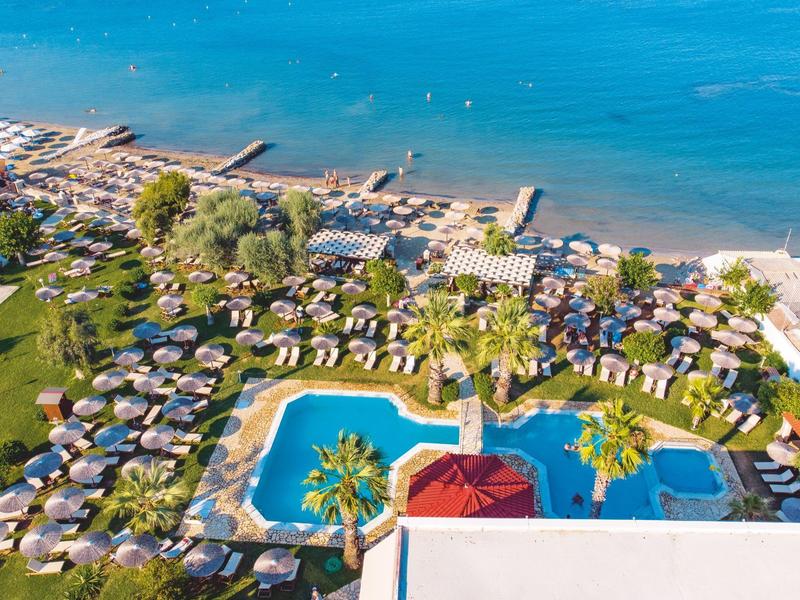 Blick auf einen Hotelpool mit Palmen und dahinter Strand mit Stränden und blauem Meer.