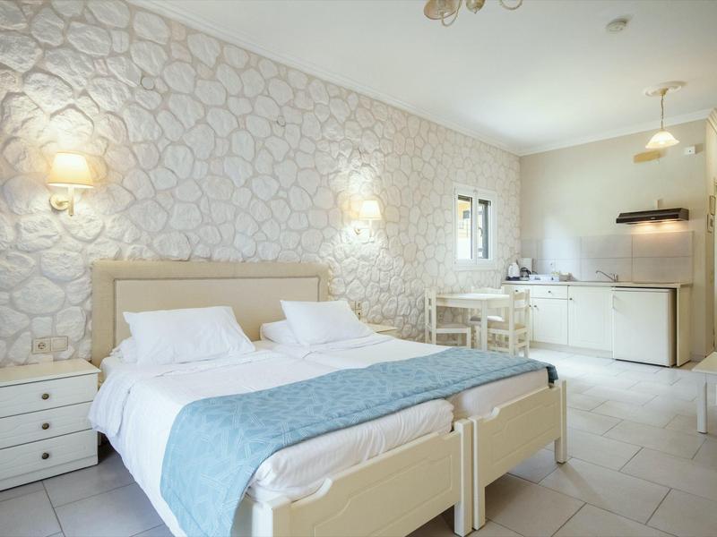 Helles Schlafzimmer mit Doppelbett, weißen Möbeln und minimalistischer Küchenzeile im Hintergrund.