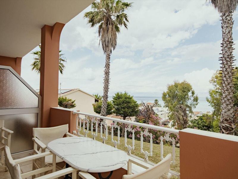 Balkon mit weißem Tisch und Stühlen, Palmen und Meer im Hintergrund bei bewölktem Himmel.