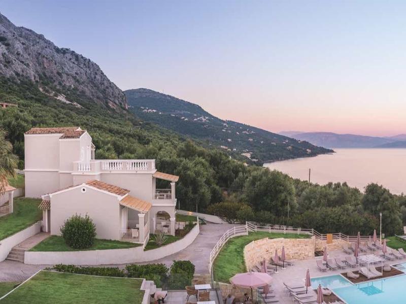 Blick auf ein Resort mit Pool, grünen Gärten, weißen Gebäuden und Meer vor Bergkulisse bei Sonnenuntergang.