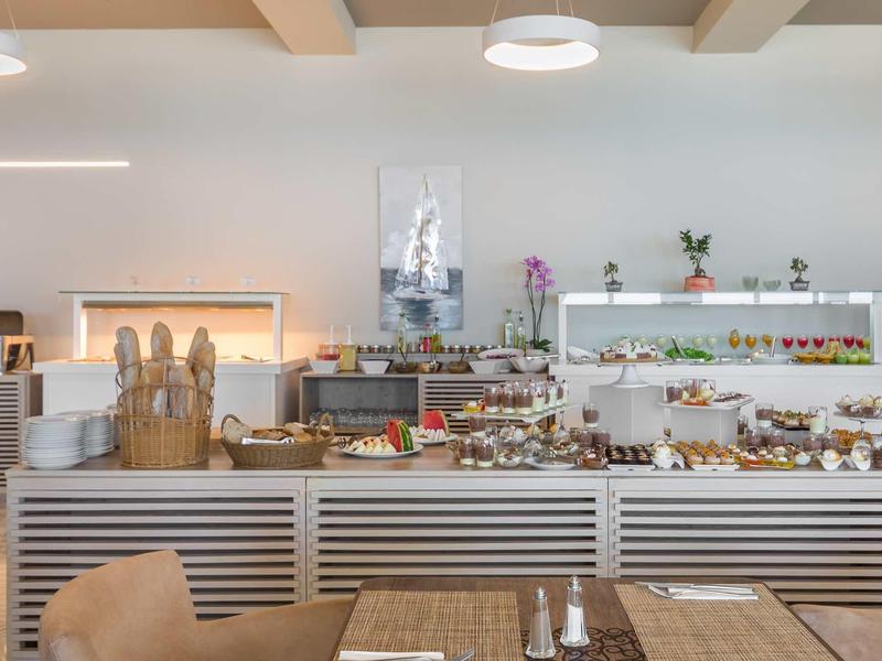 Bäckerei mit Theke voller Brote, Gebäck und Torten, helle Wände, Holztische und Stühle.