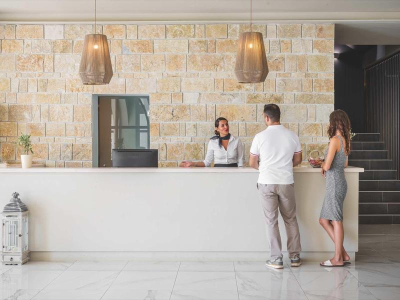 Hotelrezeption mit zwei Gästen, die am weißen Tresen mit einer Mitarbeiterin sprechen.