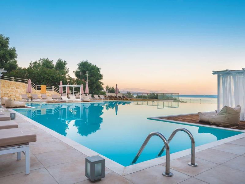 Schwimmbad mit klar blauem Wasser, weißen Liegestühlen und Blick auf ruhiges Meer bei Sonnenuntergang.