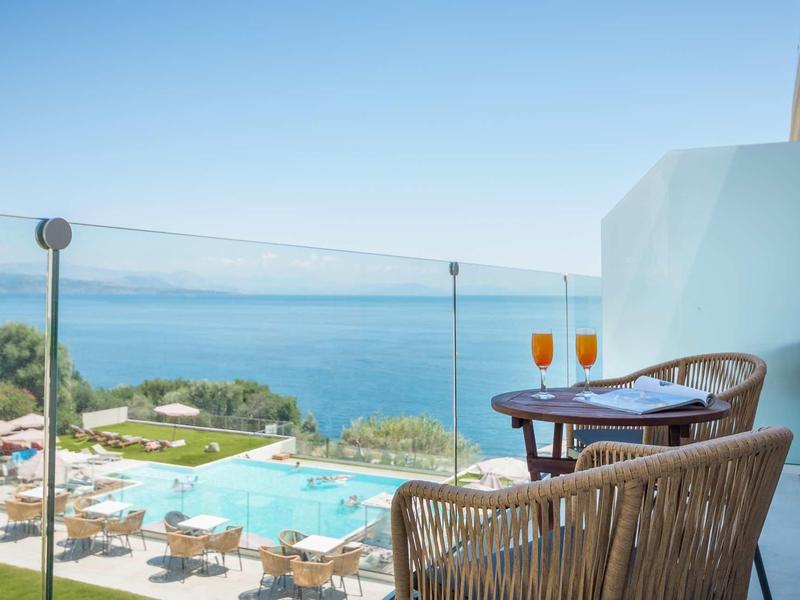 Meerblick mit Pool, Sonnenschirmen und Getränken auf einem Balkon mit Korbstühlen.