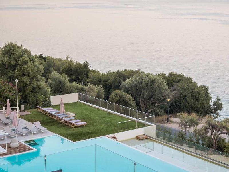 Moderne Terrasse mit Pool, Glasgeländer, Sitzmöbeln und Blick auf ruhiges Meer und Berge im Sonnenuntergang.