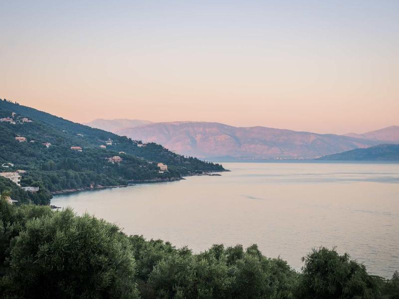 Küstenlandschaft mit Hügeln, Vegetation im Vordergrund und ruhigem Wasser bei Sonnenuntergang.
