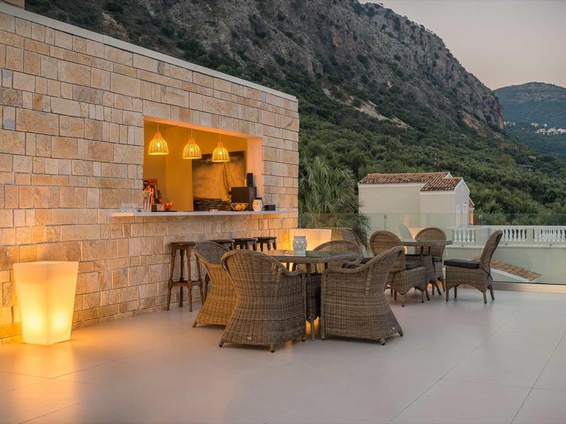 Terrasse mit Sitzgruppe und beleuchtetem Außenbarbereich vor Berglandschaft bei Dämmerung.