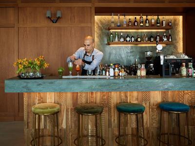 Barman przygotowujący drinki przy nowoczesnym barze z zielonym blatem i stołkami.