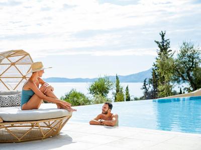 Frau sitzt auf Rattansessel am Pool, Mann entspannt im Wasser, Hintergrund Meer und Hügel.
