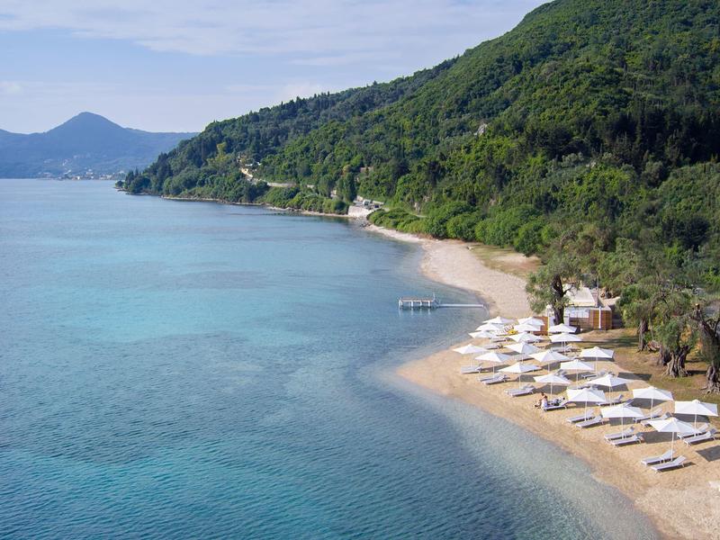 Plaża z leżakami i parasolami obok pagórkowatego, zalesionego wybrzeża i czystej niebieskiej wody.