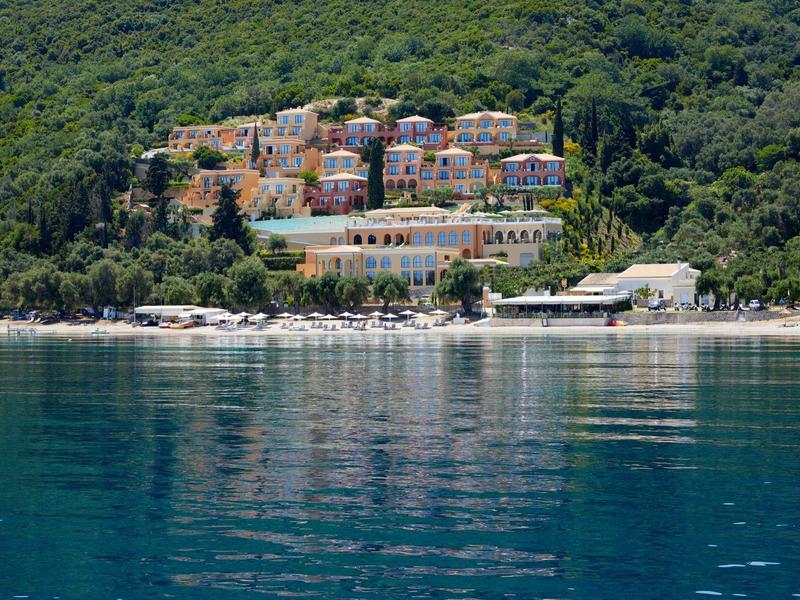 Ośrodek hotelowy na skraju lasu z widokiem na spokojną, niebieską wodę i plażę z leżakami.