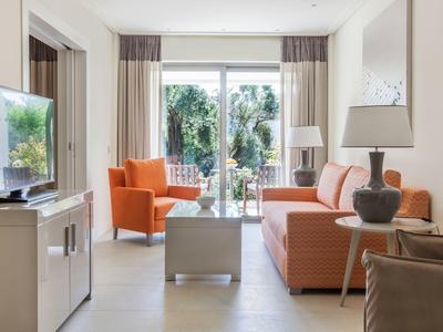 Helles Wohnzimmer mit orangem Sofa, Sessel, Glastisch und großen Fenstern mit Blick ins Grüne.