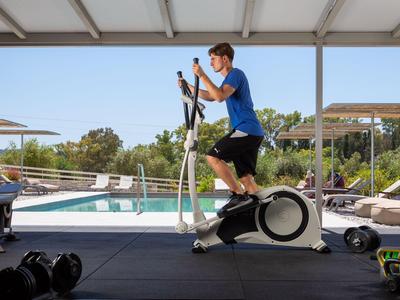 Žena cvičí na eliptickém trenažéru venku s bazénem a slunečníky v pozadí.