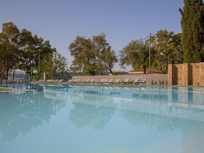 Velký venkovní bazén s lehátky a stromy v pozadí pod jasnou oblohou.