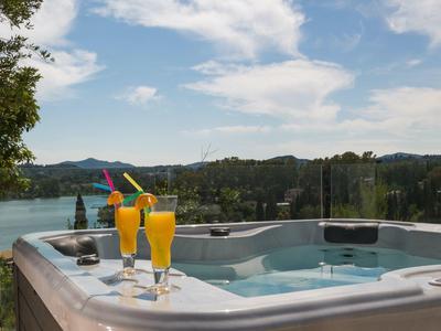 Whirlpool auf Terrasse mit vier orangenen Getränken vor Landschaft aus Wald und See unter blauem Himmel.