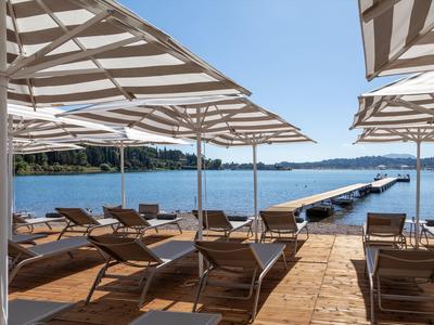 Sonnenschirme und Liegestühle auf Holzterrasse am See mit Steg und bewaldetem Ufer.