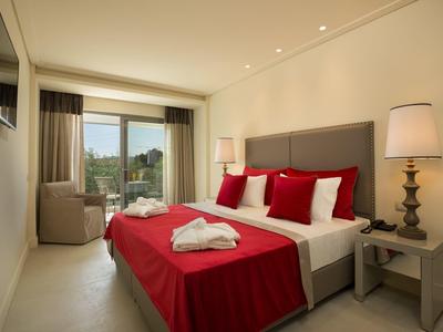 Schlichtes, helles Hotelzimmer mit großem Doppelbett, rotem Bettüberwurf und Blick auf Balkon.