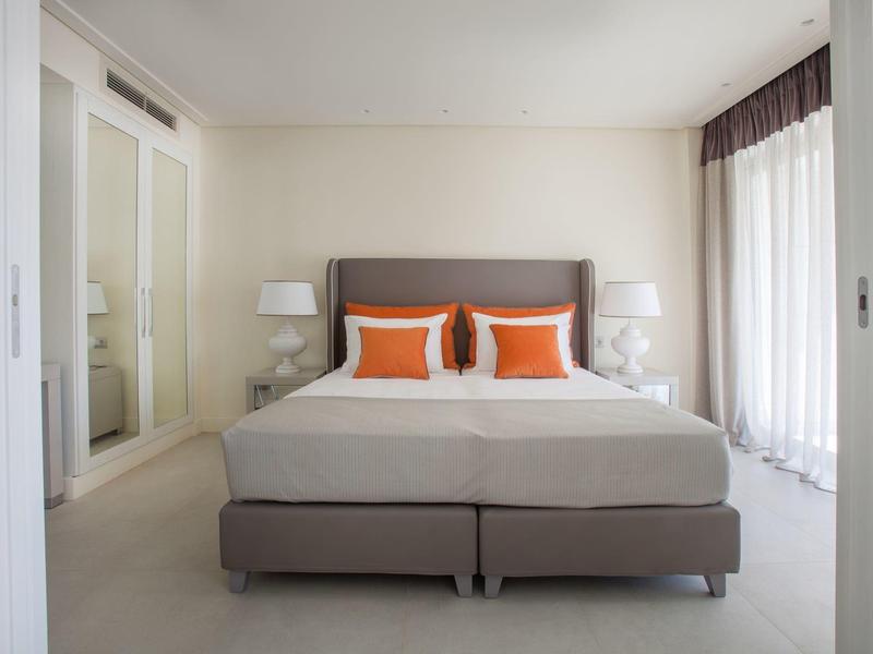 Modernes Schlafzimmer mit grauem Bett, orangen Kissen, weißen Wänden und zwei Nachttischen mit Lampen.