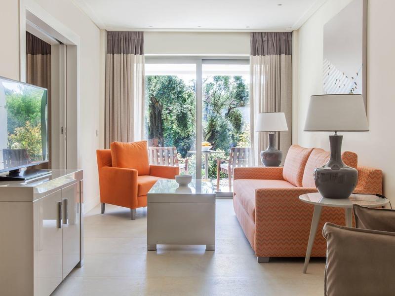 Helles Wohnzimmer mit orangem Sofa, Sessel, Glastisch und großen Fenstern mit Blick ins Grüne.