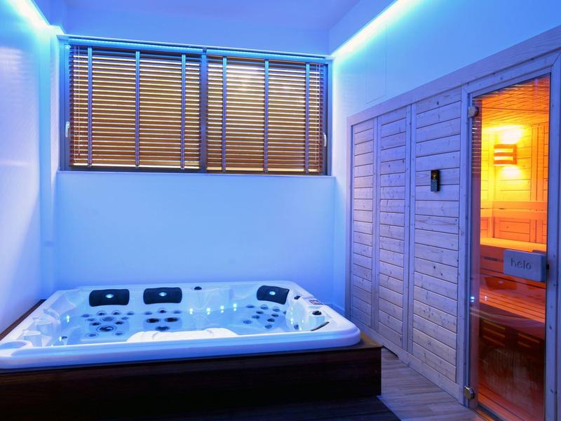 Moderner Wellnessraum mit beleuchtetem Whirlpool und Sauna, helle Wände, Holzjalousien am Fenster.