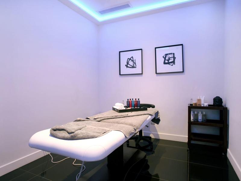 Heller Behandlungsraum mit Massageliege, zwei Bilder, kleiner schwarzer Schrank und sanfter Deckenbeleuchtung.